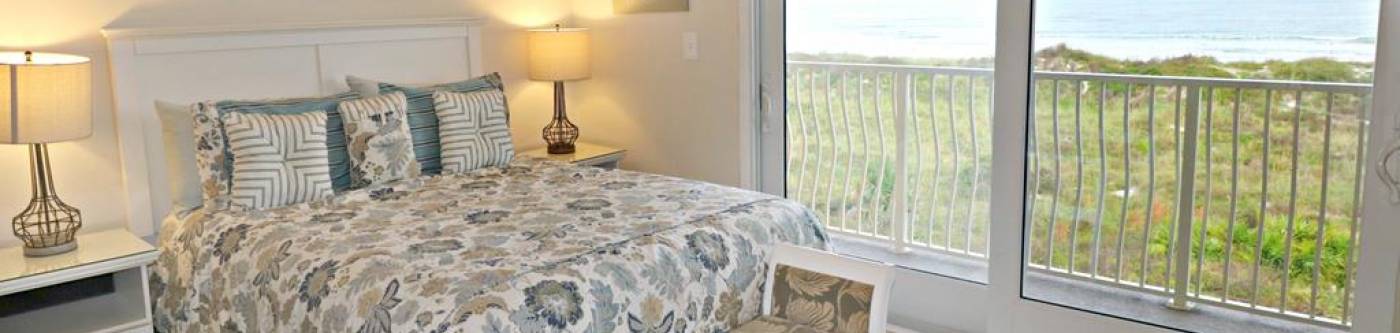 neutral bedding with light green floral print in neutral room with floor to ceiling windows overlooking dunes and beaches of St Augustine from Creston House condo rental